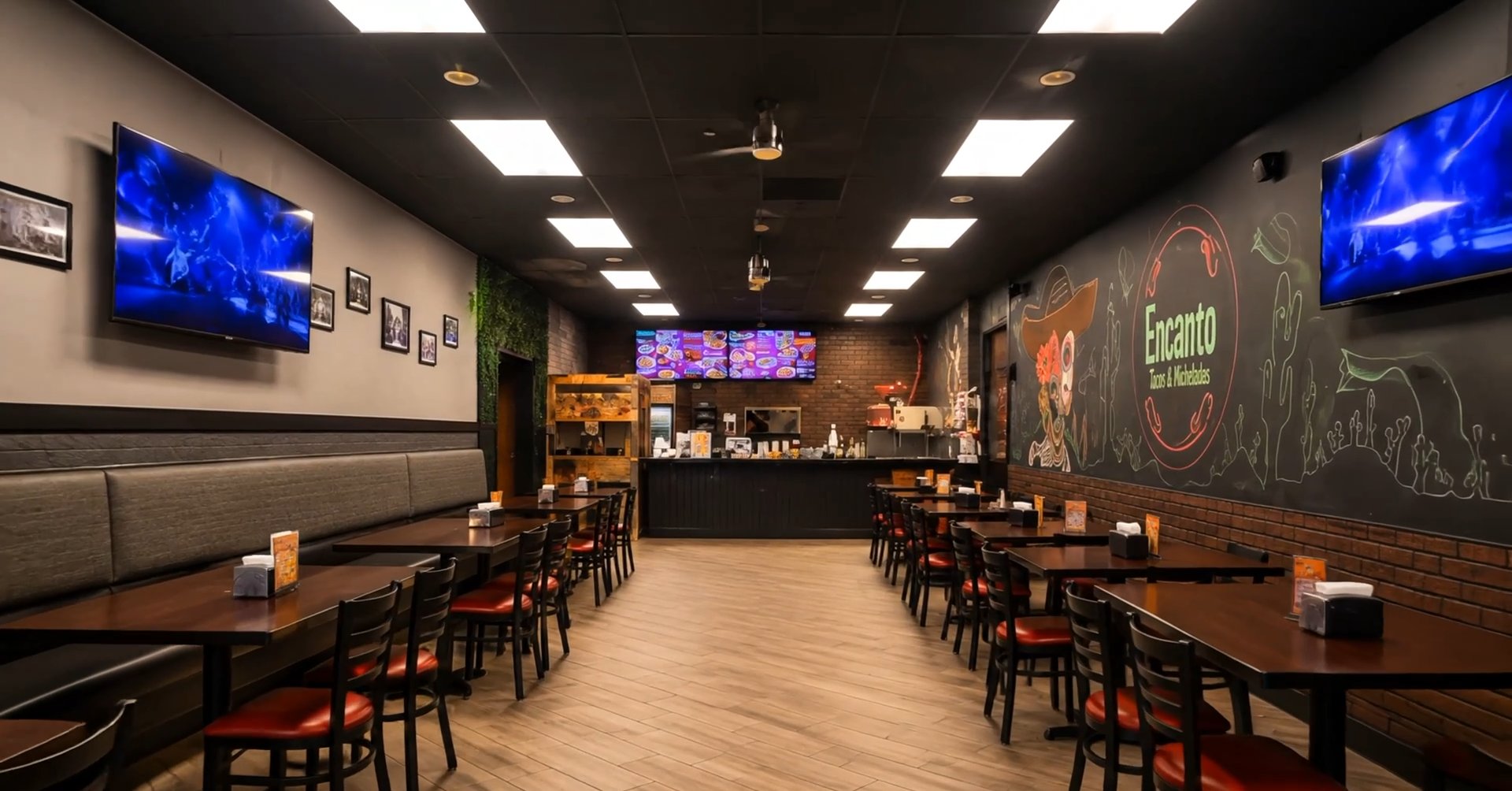 Encanto's dining room — dark ceiling with warm panel lights, wood floors, red-cushioned chairs, TVs, and the chalk-art mural leading to the service counter